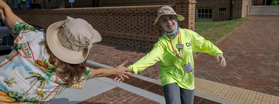 View of the back of President Rowe on a golf cart high-fiving a smiling orientation aide as they pass by on campus
