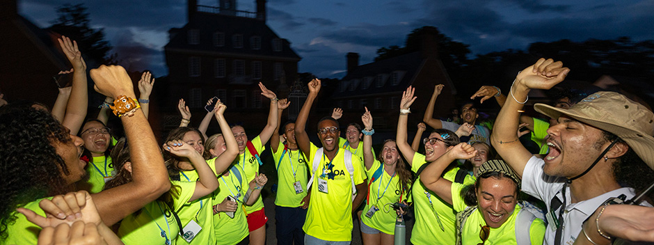 A large group of cheering orientation aides at night with Colonial Williamsburg silhouetted in the background