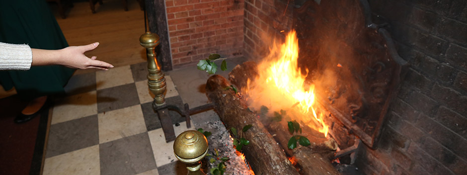 A hand tossing a sprig of holly into a roaring fire in the Great Hall fireplace